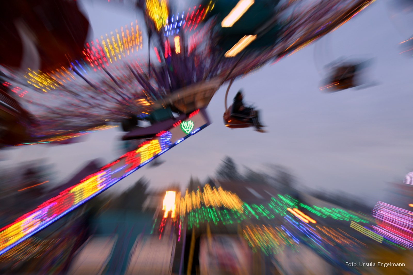 Fahrgeschäft in voller Fahrt mit bunten Lichtspuren in der Abenddämmerung, fotografiert aus der Bewegung – Stimmungsbild, Foto: Ursula Engelmann